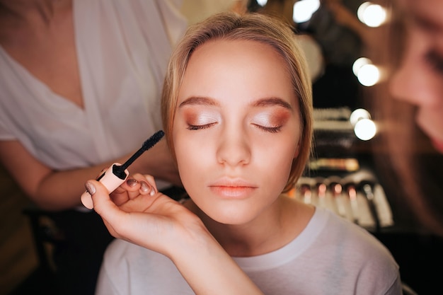 Schöne junge blonde Frau Gesicht hautnah. Sie hat Lidschatten auf der Lidlinie. Maskenbildner verwenden Mascara. Friseur Arbeit hinter.