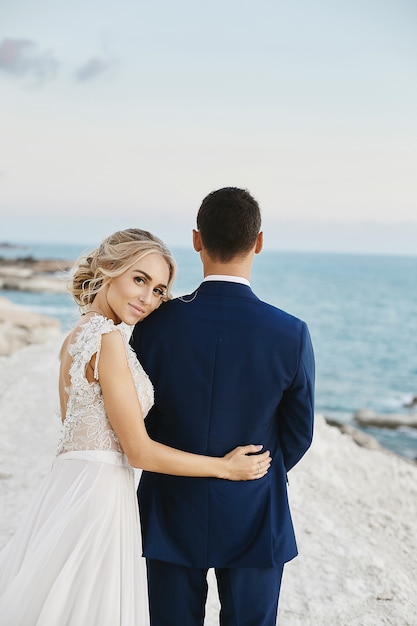 Schöne junge blonde Frau im weißen Spitzenkleid stützt sich auf den gutaussehenden Mann im stilvollen blauen Anzug auf dem weißen Felsen an der Küste der Adria. Modell Mädchen in einem Hochzeitskleid mit stilvollem Bräutigam