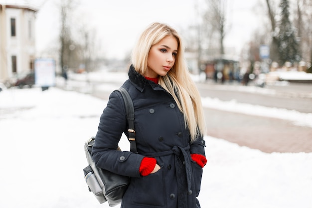 Schöne junge blonde Frau in einem Modewintermantel mit einer stilvollen Tasche geht in die Stadt