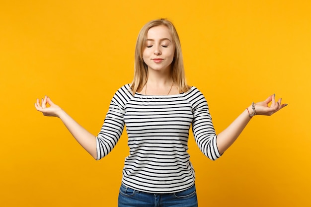 Schöne junge blonde Frau in gestreifter Kleidung hält die Hände in Yoga-Geste, isoliert auf leuchtend gelb-orangefarbenem Hintergrund, Studioportrait. Menschen aufrichtige Emotionen Lifestyle-Konzept. Mock-up-Kopienbereich.