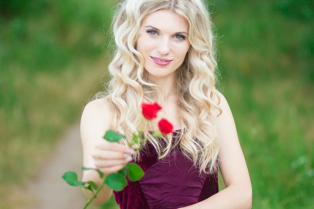 Schöne junge blonde Frau mit dem lockigen Haar, das einen Korb voll von den roten Rosen anhält. Weicher Fokus