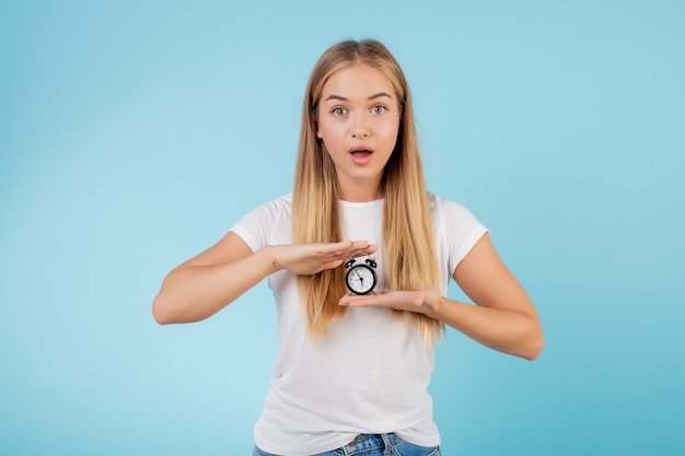 Foto schöne junge blonde frau mit der alarmuhr, welche die zeit getrennt über blau zeigt