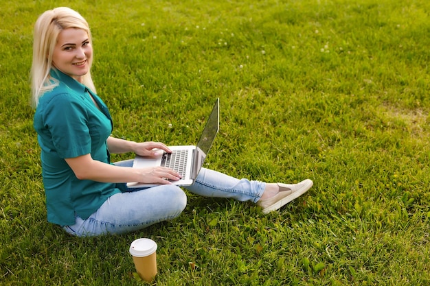 Schöne junge blonde Frau mit einem Laptop im Park an einem warmen Sommertag