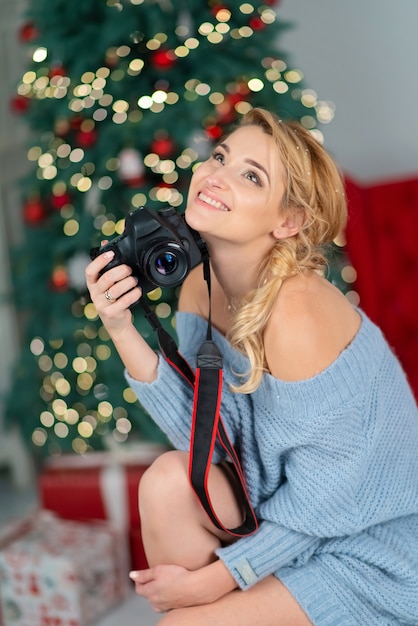 Schöne junge blonde Frau mit einer Fotokamera lächelt neben einem Weihnachtsbaum Bokeh.