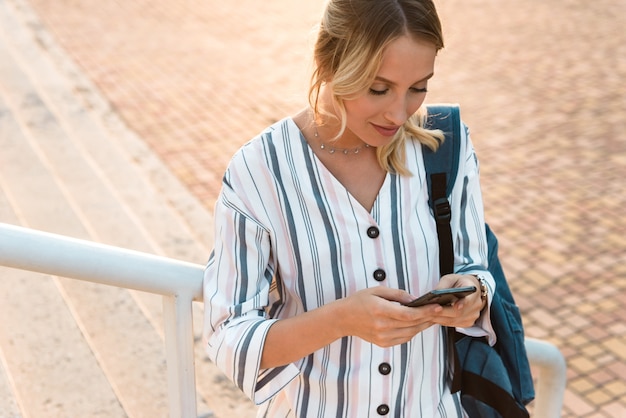 Foto schöne junge blonde frau mit rucksack mit handy beim stehen auf stufen im freien