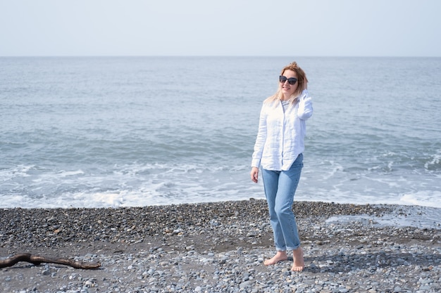 Schöne junge blonde Frau mit Sonnenbrille, die am Ufer des Meeres steht und lächelnd in die Kamera schaut