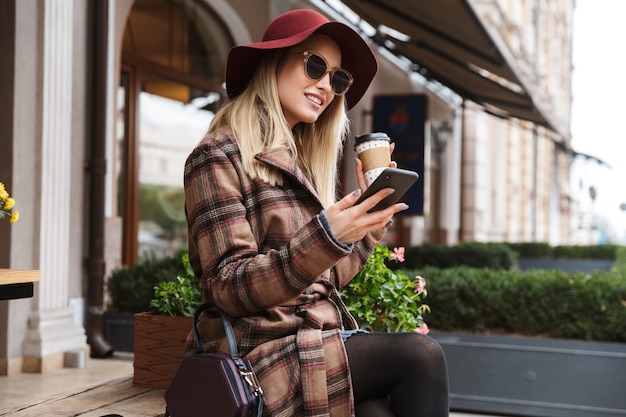 Schöne junge blonde stilvolle Frau, die einen Mantel trägt, der draußen, unter Verwendung des Mobiltelefons sich entspannt