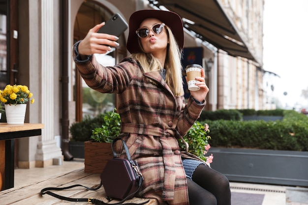 Schöne junge blonde stilvolle Frau, die einen Mantel trägt, der sich im Freien entspannt und ein Selfie nimmt