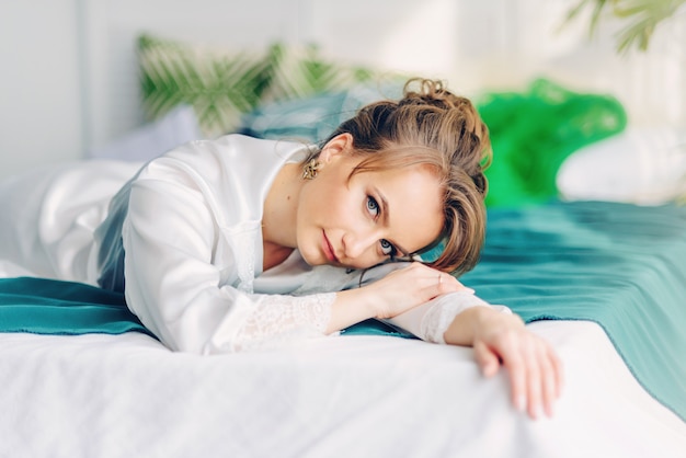 Schöne junge Braut, die auf dem Bett liegt, bedeckt mit einer grünen Decke in einem Raum mit Blumen