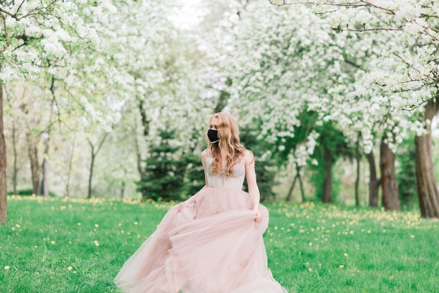 Schöne junge Braut in einem Hochzeitskleid und einer schwarzen medizinischen Maske auf ihrem Gesicht nahe einem blühenden Apfelbaum. Covid19 Schutz.