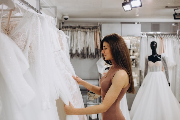 Schöne junge Braut schaut in den Spiegel und lächelt, während sie elegante Brautkleider im modernen Hochzeitssalon auswählt