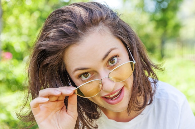 Schöne junge brünette Frau in der trendigen gelben Sonnenbrille, die in einem Park oder im Garten ruht und lächelt