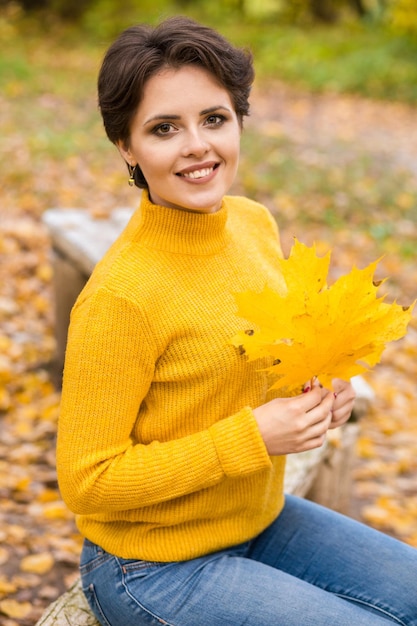 Schöne junge brünette Frau in einem gelben Strickpullover posiert in einem Herbstpark mit einem Strauß Blätter