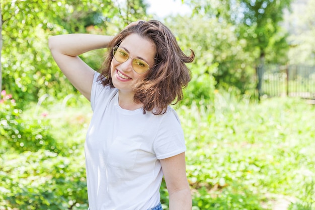 Schöne junge Brünette Frau mit braunen Haaren in der trendigen gelben Sonnenbrille, die auf grünem Hintergrund des Parks oder des Gartens ruht und lächelt.