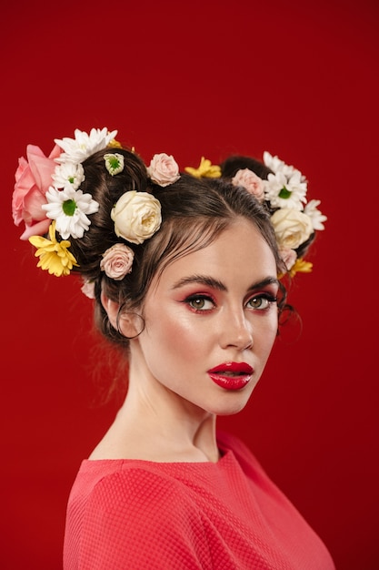 Schöne junge brünette Frau mit einer erstaunlichen Blumenfrisur mit Make-up, die isoliert über roter Wand posiert und nach vorne schaut