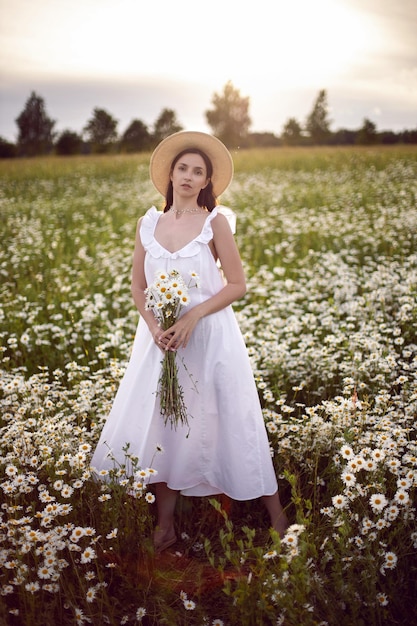 Schöne junge brünette Frau mit langen Haaren in einem Hut und einem weißen Kleid hält einen Blumenstrauß in ihren Händen, der im Sommer bei Sonnenuntergang auf einem Kamillenfeld steht