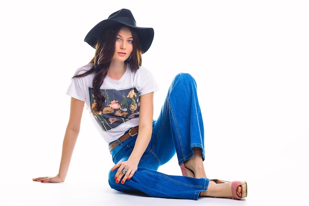 Schöne junge Brünette in Jeans Glockenboden weißes T-Shirt und schwarzer Hut, auf dem Boden sitzend