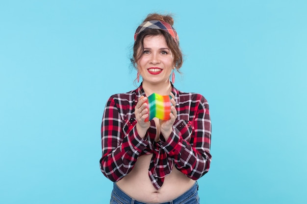 Schöne junge coole Frau auf einem karierten Hemd, das einen Spielzeugregenbogen in ihren Händen hält, der auf einem blauen Hintergrund aufwirft.