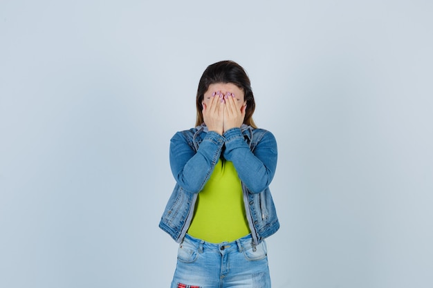 Schöne junge Dame, die Gesicht mit den Händen in Denim-Outfit bedeckt und beleidigt aussieht, Vorderansicht.