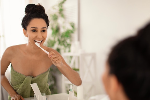 Schöne junge Dame im Handtuch, die Zähne mit Zahnbürste in der Nähe des Spiegels zu Hause putzt, Kopienraum zahnärztlich