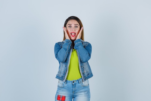 Schöne junge Dame in Denim-Outfit, die die Hände in der Nähe des Gesichts hält und aufgeregt aussieht, Vorderansicht.