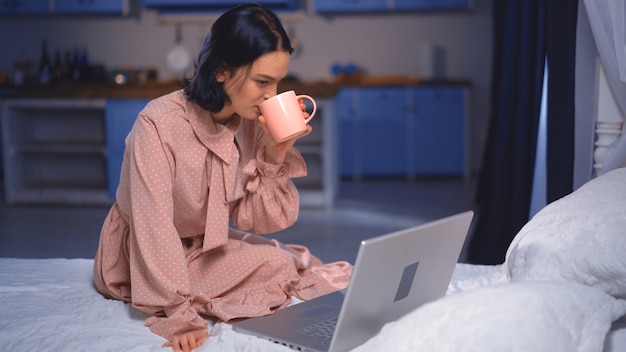 Schöne junge dunkelhaarige Frau, die auf dem Bett sitzt und eine Tasse Kaffee schmeckt