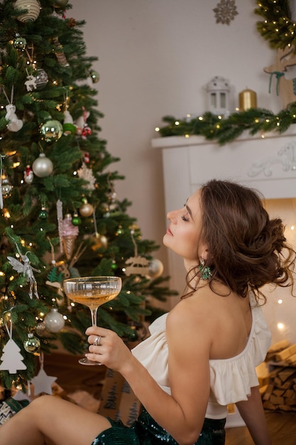 Schöne junge dunkelhaarige Frau in stilvollen Kleidern in der Nähe des Weihnachtsbaums mit einem Glas Champagner Feiertage Neujahr
