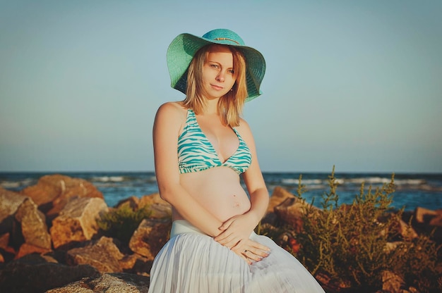 Schöne junge elegante schwangere blonde Frau in einem blauen Hut geht am Strand, Küste bei Sonnenuntergang.
