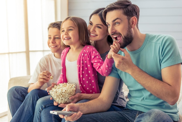 Schöne junge Eltern, ihre Tochter und ihr Sohn sehen fern, essen Popcorn und lächeln, sitzen zu Hause auf dem Sofa. Papa benutzt eine Fernbedienung