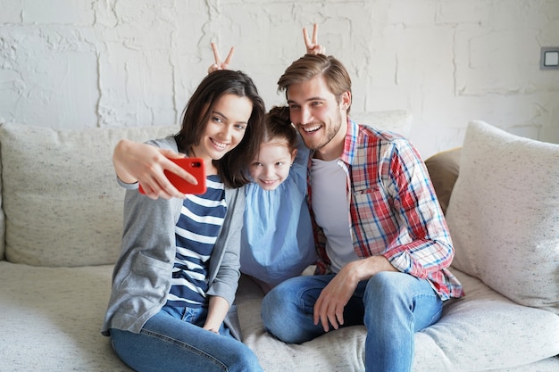 Schöne junge Familie mit kleinem Kind, das zu Hause auf der Couch ein Selfie mit einem Smartphone macht.