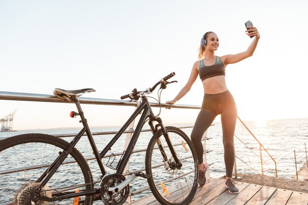 Schöne junge Fitness-Radfahrerin in Sportkleidung, die mit dem Fahrrad am Pier steht und ein Selfie macht