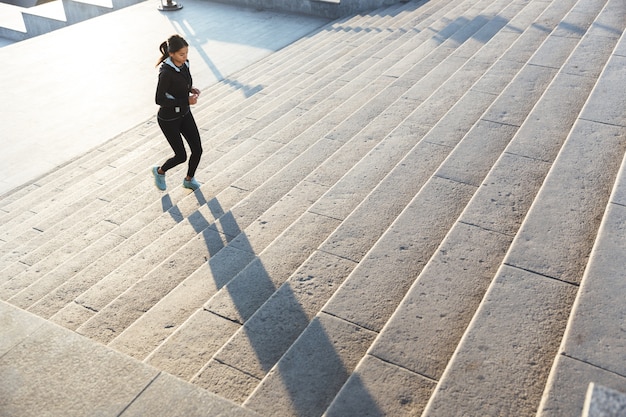 Schöne junge Fitnessfrau, die Sportkleidung trägt, die an der Treppe draußen joggt