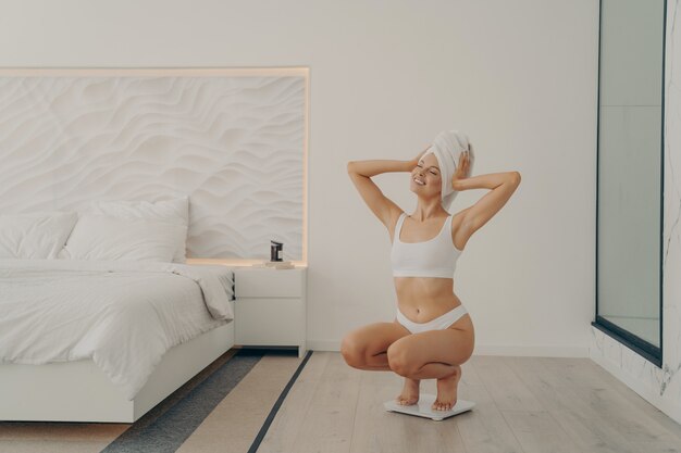 Schöne junge, fitte weibliche Model hockt auf Waagen im modernen Schlafzimmerinterieur nach der morgendlichen Duschroutine in Unterwäsche glücklich und zufrieden mit dem Ergebnis ihres Gewichts. Gewichtsverlust und Diätkonzept
