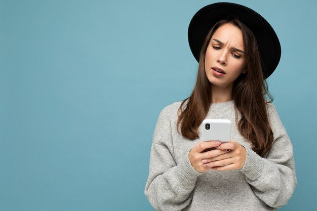 Schöne junge fragende unzufriedene brünette Frau mit schwarzem Hut und grauem Pullover mit Smartphone, die isoliert auf Hintergrund schaut. Platz kopieren