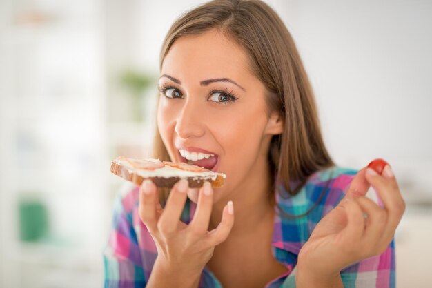 Schöne junge Frau beim Frühstück in der heimischen Küche. Sie isst ein frisches Sandwich und trinkt Milch. Blick in die Kamera.