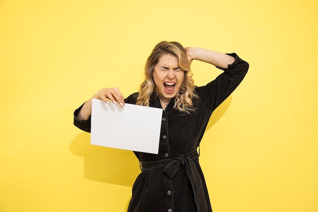 Schöne junge Frau. Blondes Mädchen schreit und hält den Kopf mit der Hand. hält leeres Papier A4 für Handelszeichen, negative Emotionen. Foto auf gelbem Hintergrund.