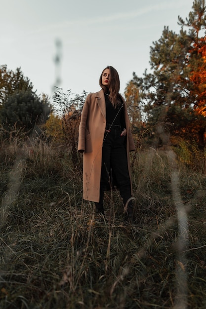 Schöne junge Frau der Mode in einem beige Mantel, in einem schwarzen Pullover und in einer Jeans auf einer Natur auf einem Feld mit Gras