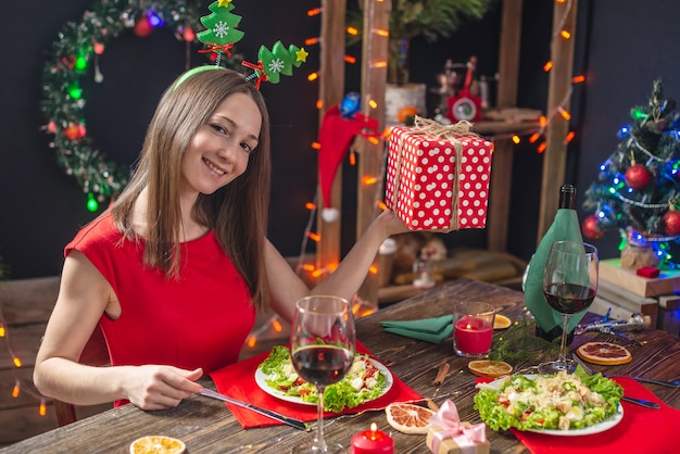 Schöne junge Frau, die am Feiertagstisch mit roter Geschenkbox in Weihnachtsdekorationen speist