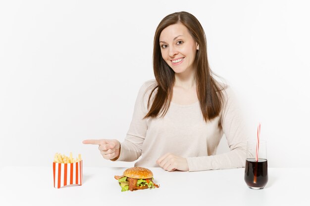 Schöne junge Frau, die am Tisch mit Burger, Pommes-Frites, Cola in der Glasflasche lokalisiert auf weißem Hintergrund sitzt. Richtige Ernährung oder amerikanisches klassisches Fast Food. Werbefläche mit Kopienraum.