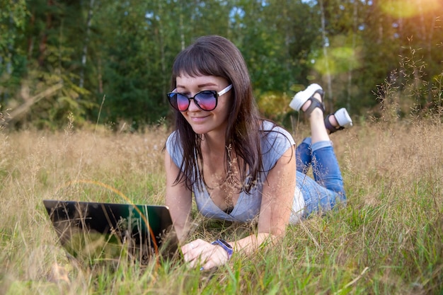 Schöne junge Frau, die an einem warmen Sommertag auf einem sonnigen Rasen emotional an einem Laptop arbeitet. glücklicher Remote-Freiberufler