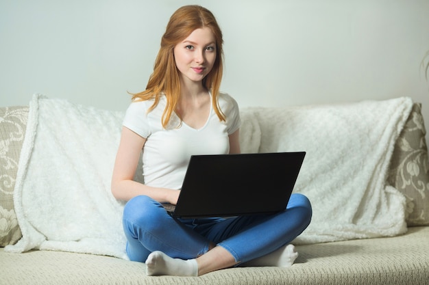 schöne junge Frau, die auf dem Sofa mit einem Laptop sitzt