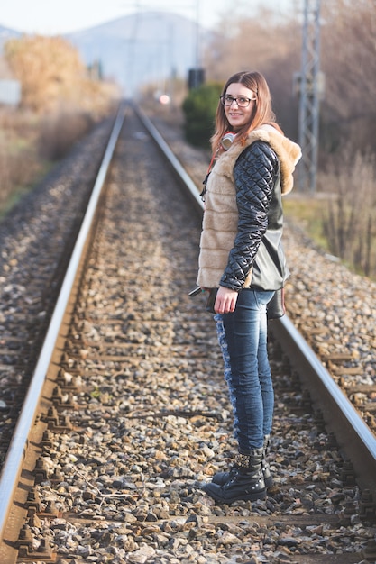 Schöne junge Frau, die auf Eisenbahnlinien steht. Die Eisenbahn befindet sich in einem Wohngebiet. Das Mädchen hat einen lässigen Look.