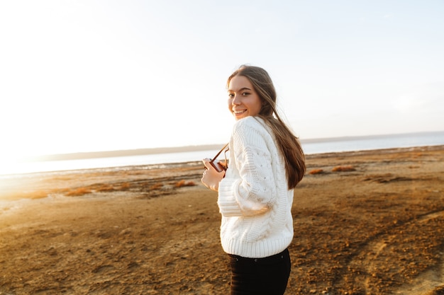 Schöne junge Frau, die draußen am Strand während des Sonnenuntergangs geht und Fotos mit Fotokamera macht