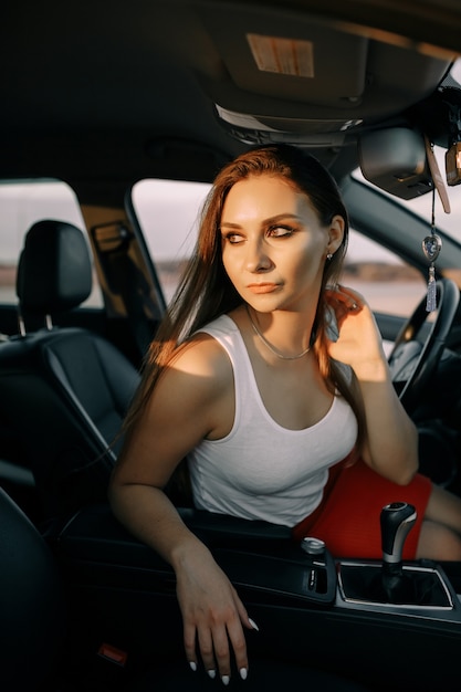 Schöne junge Frau, die ein Auto am Abend in der Sonnenuntergangssonne auf einem leeren Parkplatz fährt