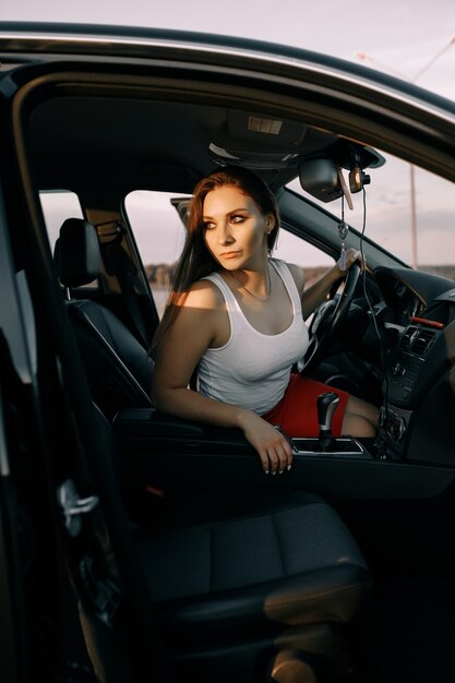 Schöne junge Frau, die ein Auto am Abend in der Sonnenuntergangssonne auf einem leeren Parkplatz fährt