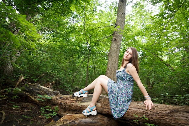Schöne junge Frau, die ein elegantes weißes Kleid trägt und auf einem Waldweg spazieren geht, während Sonnenstrahlen durch die Blätter der Bäume strahlen