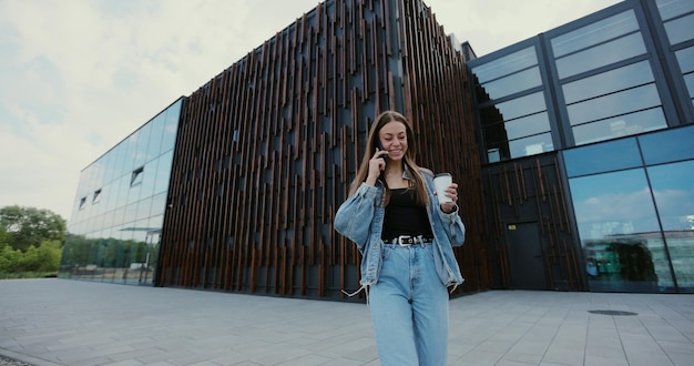 Schöne junge Frau, die eine Jeansjacke trägt und ein modernes Smartphone benutzt, während sie in der Stadt spazieren geht Frau, die mit Freunden telefoniert, die sich freuen, lachen und Kaffee trinken