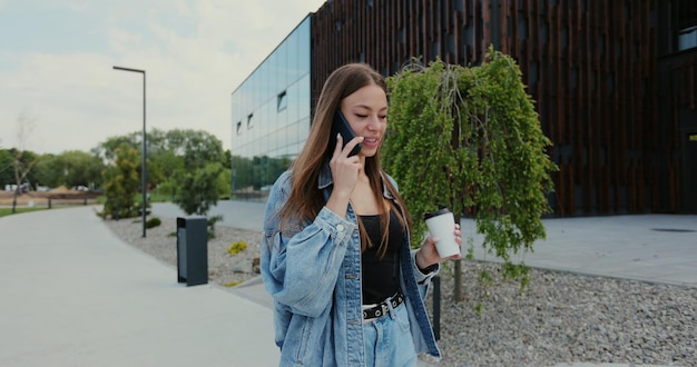 Schöne junge Frau, die eine Jeansjacke trägt und ein modernes Smartphone benutzt, während sie in der Stadt spazieren geht Frau, die mit Freunden telefoniert, die sich freuen, lachen und Kaffee trinken