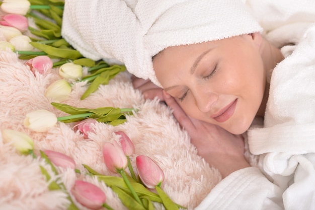 Schöne junge Frau, die einen weißen Bademantel trägt und auf dem Bett mit Blumen schläft