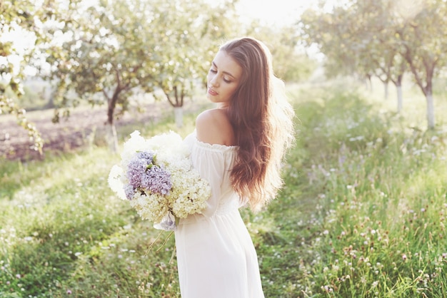 Schöne junge Frau, die elegantes weißes Kleid trägt und schönen sonnigen Nachmittag in einem Sommergarten genießt